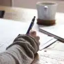 person writing on brown wooden table near white ceramic mug