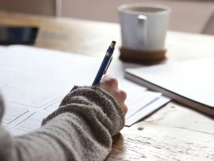 person writing on brown wooden table near white ceramic mug